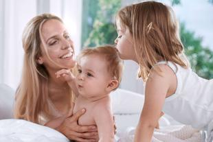 Drei Kinder sitzen auf einem Bett, eines hält ein Baby aufrecht, ein anderes beugt sich zu ihm vor