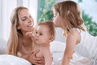 Drei Kinder sitzen auf einem Bett, eines hält ein Baby aufrecht, ein anderes beugt sich zu ihm vor