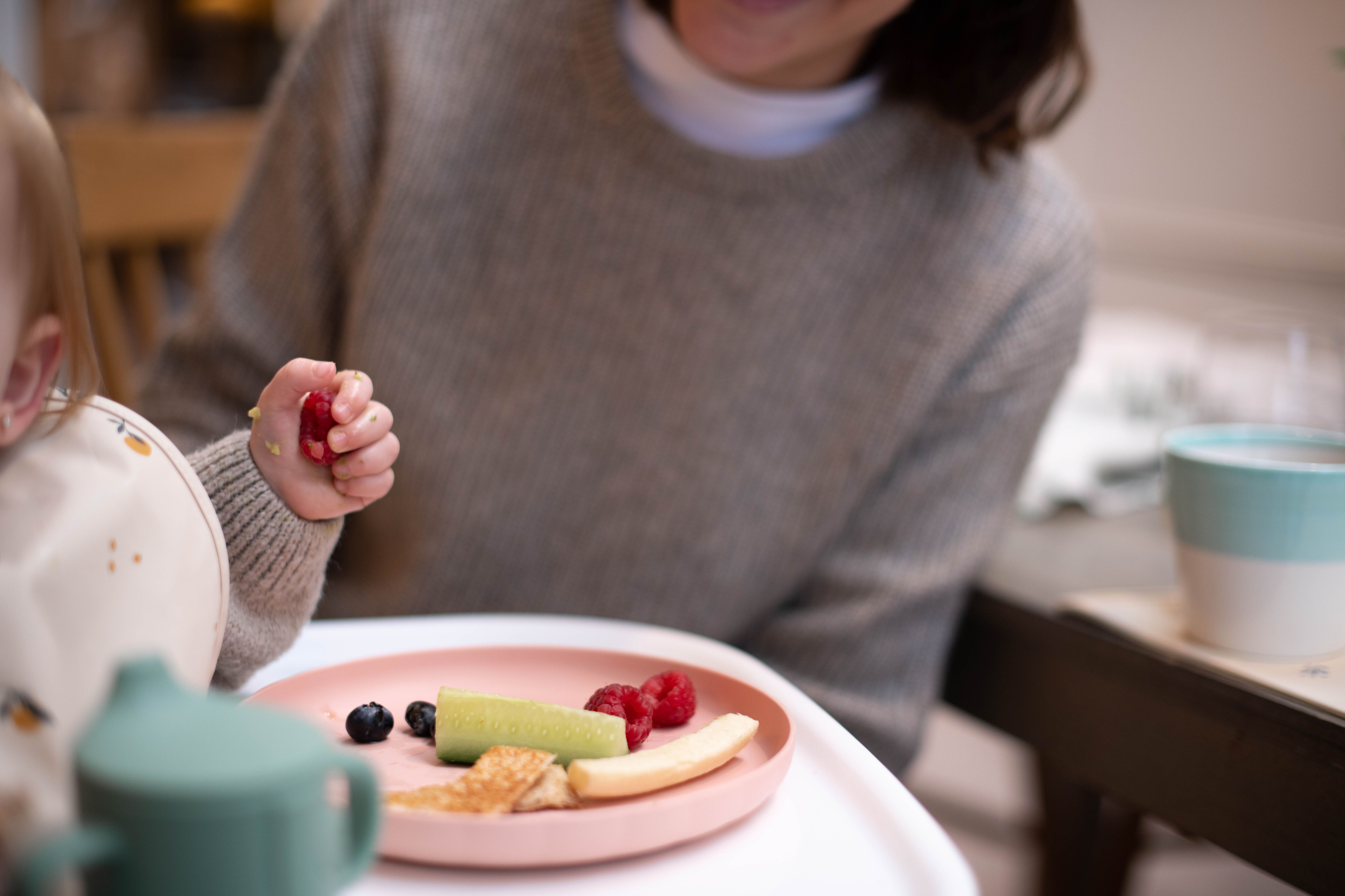 Baby practising self-feeding with finger foods on a plate while a parent sits close by, supporting calm mealtime
