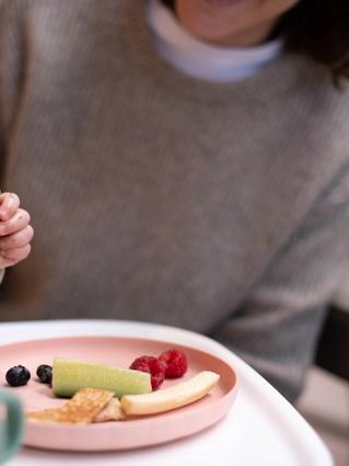 Baby practising self-feeding with finger foods on a plate while a parent sits close by, supporting calm mealtime