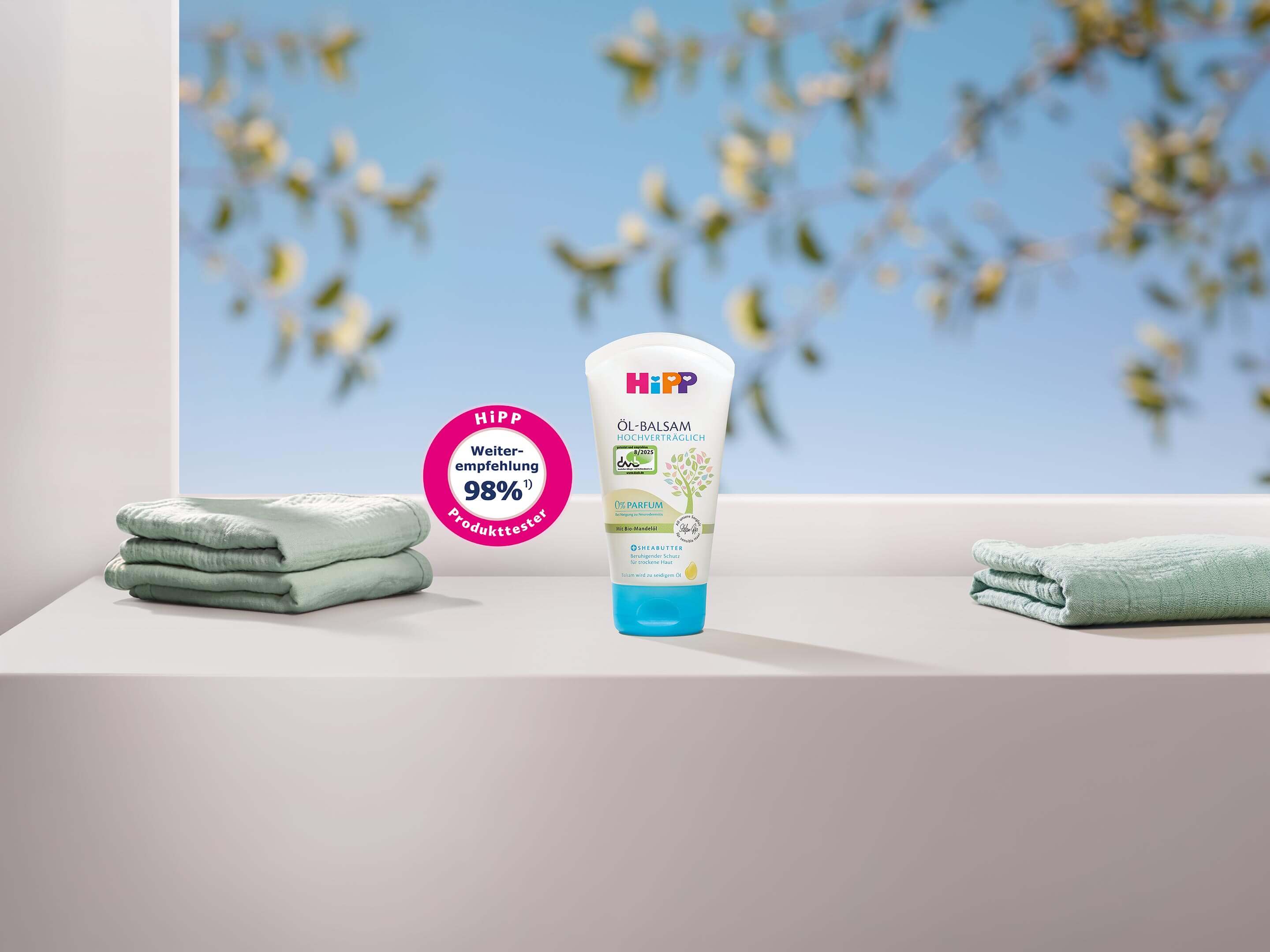 HiPP HOCHVERTRÄGLICH Öl-Balsam steht zwischen weichen Babyhandtüchern vor blauem Himmel mit sanft verschwommenen Blüten.