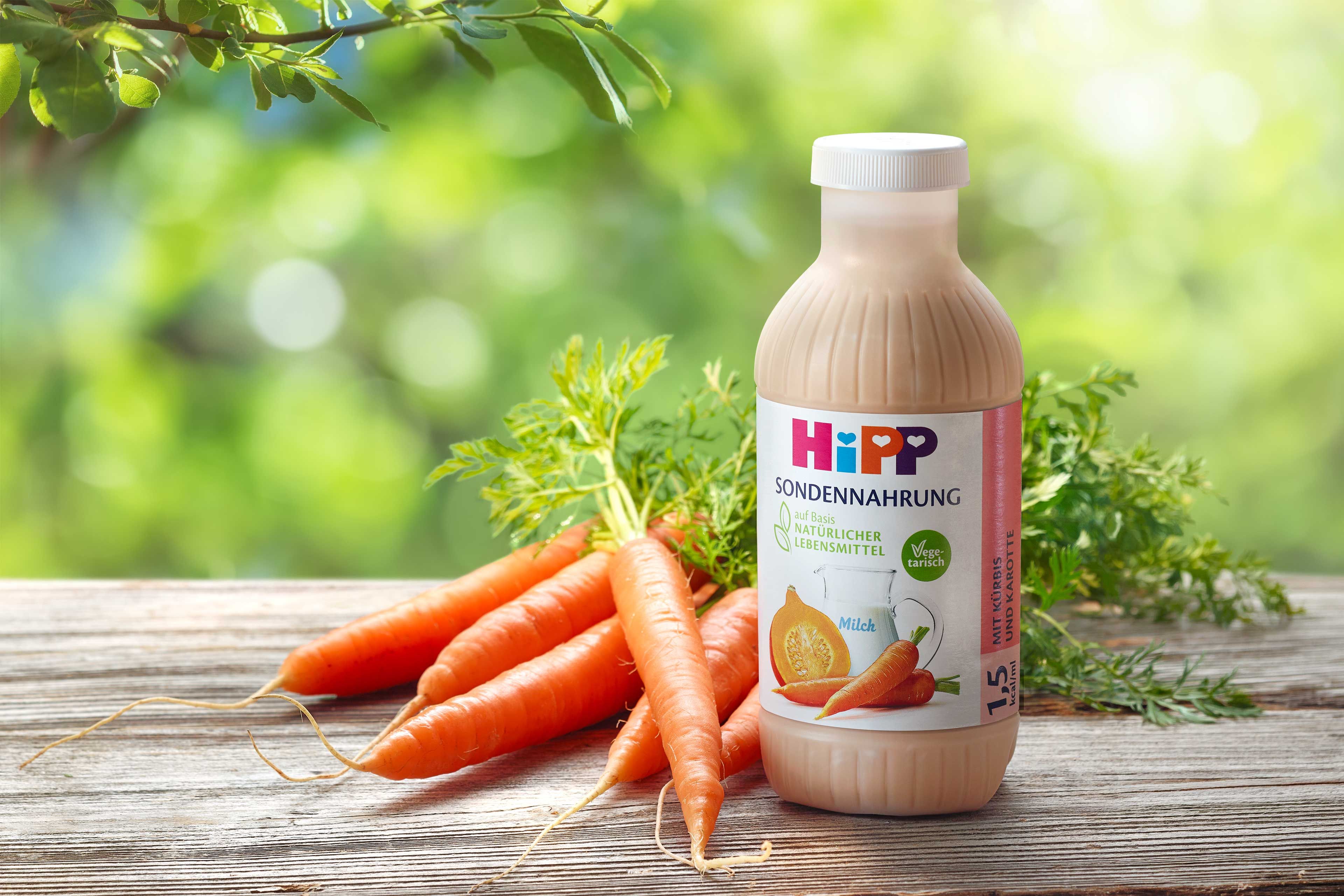 HiPP Sondennahrung Flasche mit Kürbis und Karotte steht neben frischen Karotten auf Holztisch vor grünem Naturhintergrund