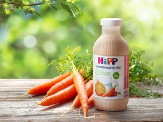 HiPP Sondennahrung Flasche mit Kürbis und Karotte steht neben frischen Karotten auf Holztisch vor grünem Naturhintergrund