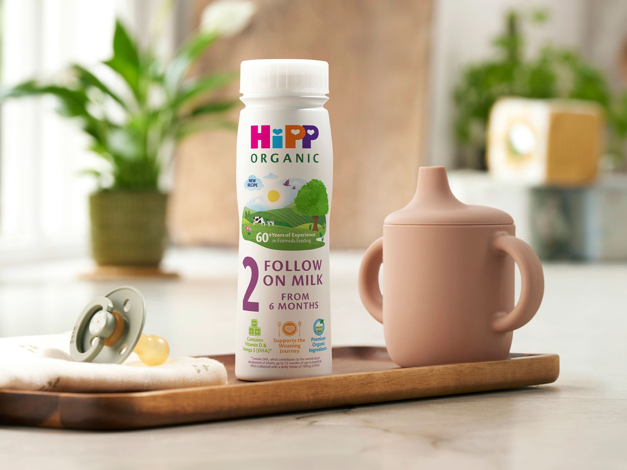 HiPP Organic follow on milk ready to feed bottle on a tray beside a pastel sippy cup and dummy on a bright kitchen worktop