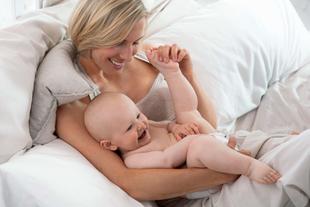 Lächelnde Mutter kuschelt im Bett mit ihrem fröhlich strampelnden Baby und genießt die gemeinsame Nähe
