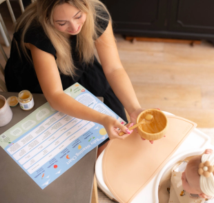 Mum feeding purée to her baby in a highchair while following a colourful baby weaning chart on the table beside her