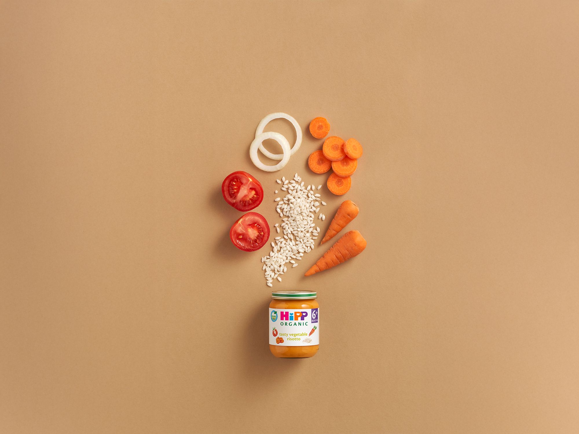HiPP Organic Tasty Vegetable Risotto jar with carrots, tomatoes, onion rings and rice arranged above on a warm beige background