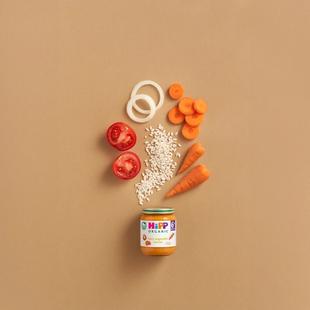 HiPP Organic Tasty Vegetable Risotto jar with carrots, tomatoes, onion rings and rice arranged above on a warm beige background