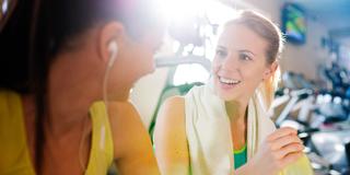 Zwei Frauen in einem Fitnessstudio, eine trägt Kopfhörer, die andere lächelt.