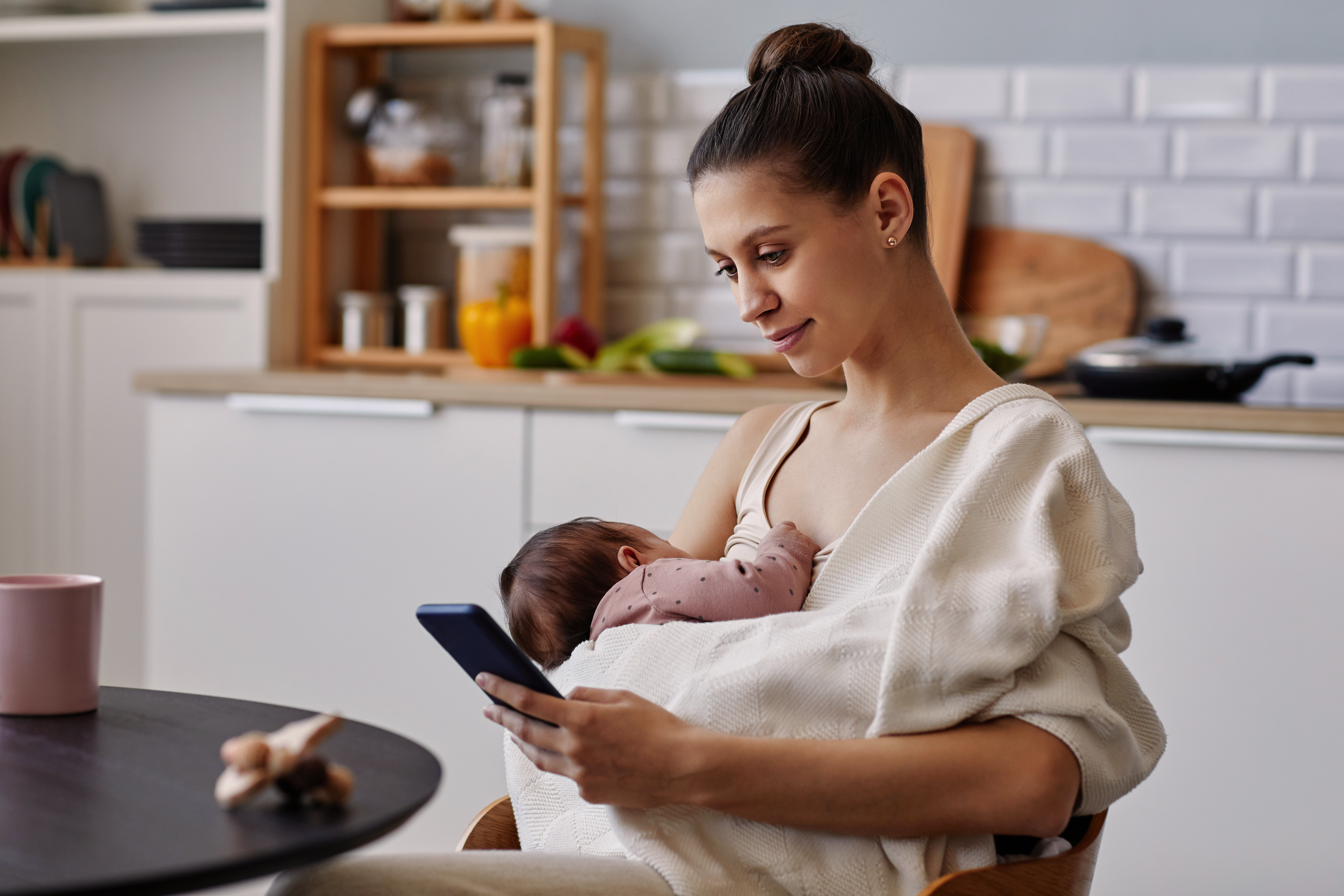 New mum breastfeeding her newborn while reading health advice on her smartphone in a calm kitchen.