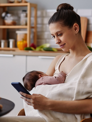 New mum breastfeeding her newborn while reading health advice on her smartphone in a calm kitchen.