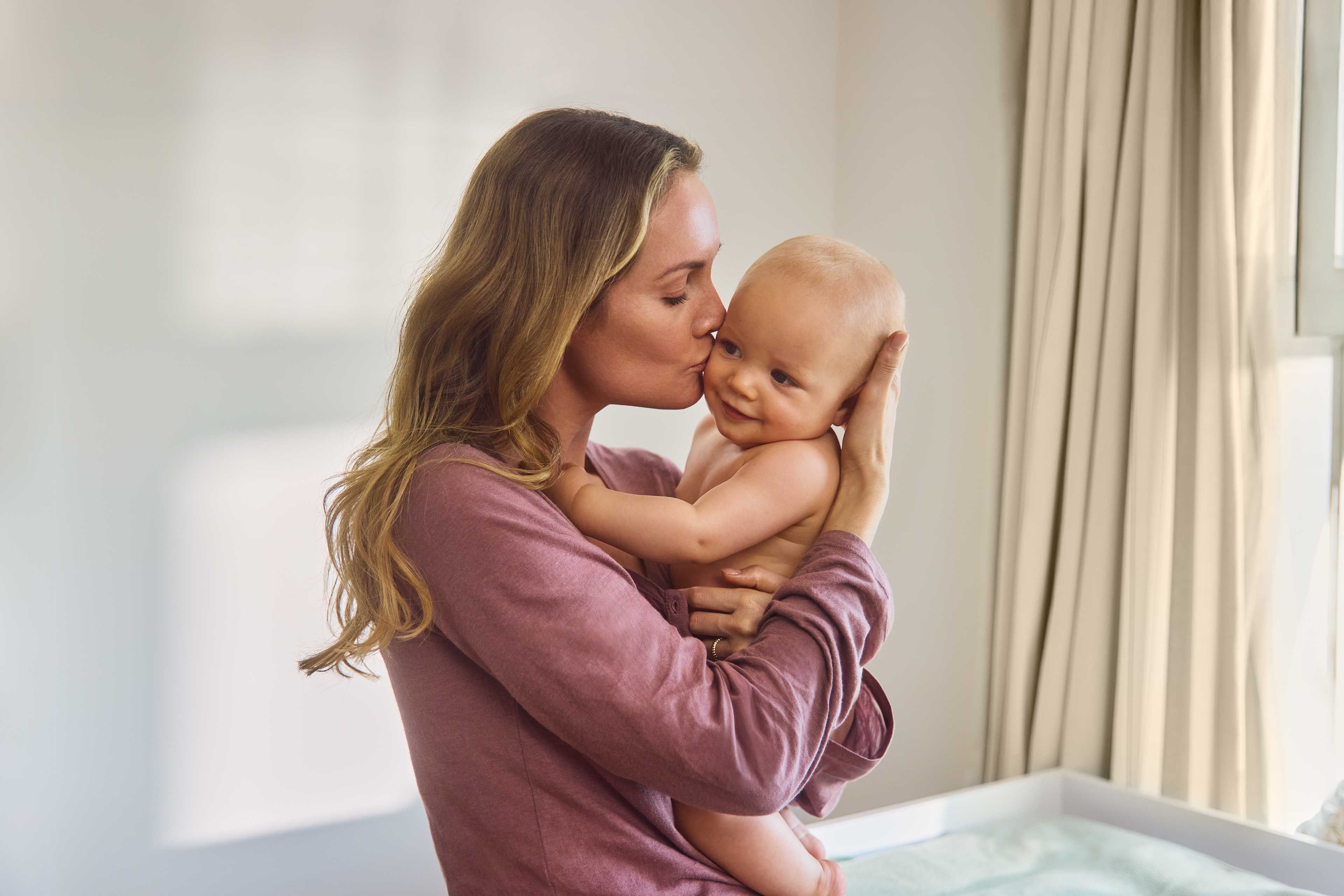 Mutter hält ihr lächelndes Baby im Arm und küsst es liebevoll auf die Wange.