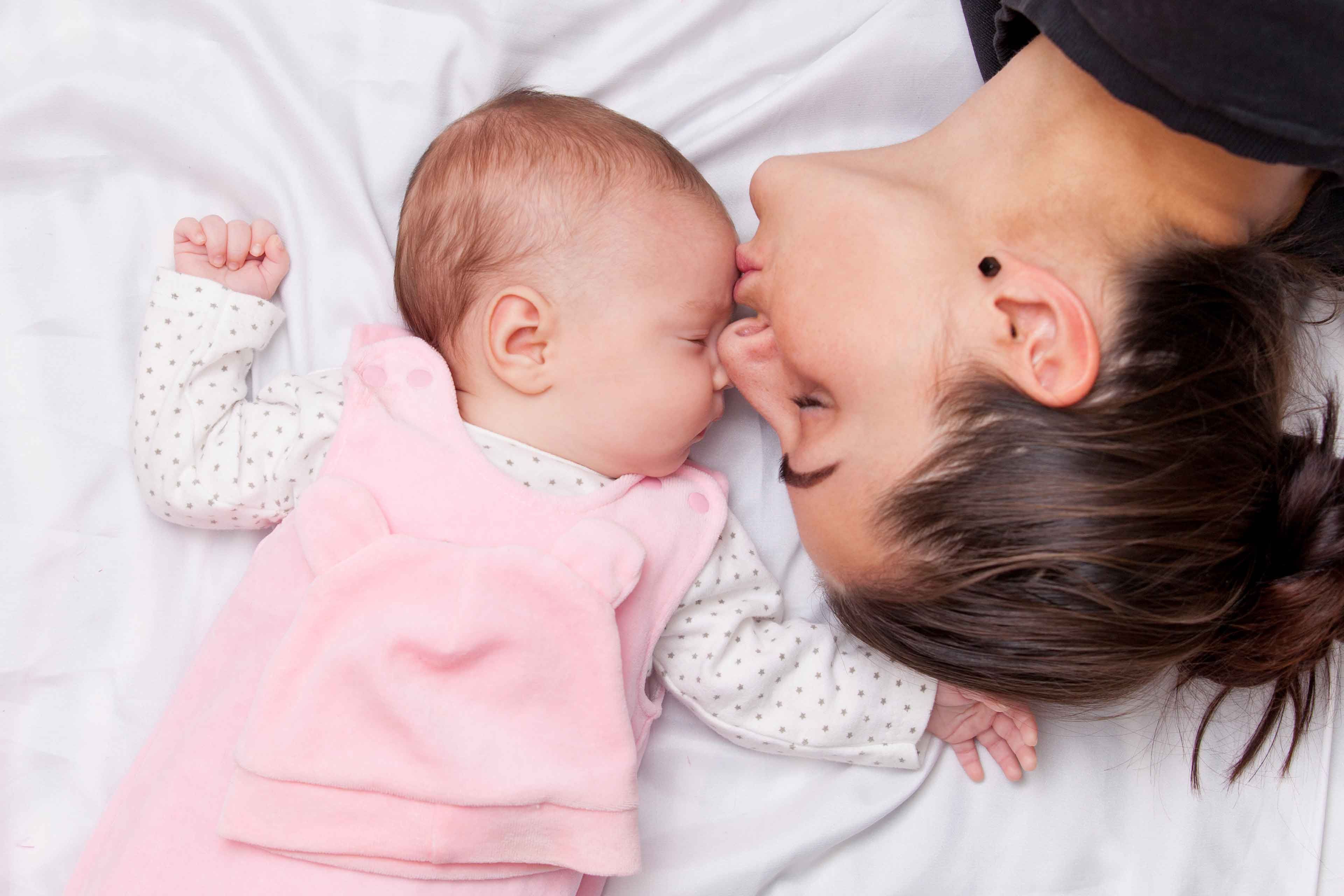 Ein zärtlicher Moment zwischen einer Mutter und ihrem Baby, in dem die Mutter sanft die Stirn des Babys küsst, während sie beide auf einem weißen Bett liegen.