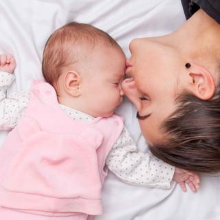 Ein zärtlicher Moment zwischen einer Mutter und ihrem Baby, in dem die Mutter sanft die Stirn des Babys küsst, während sie beide auf einem weißen Bett liegen.
