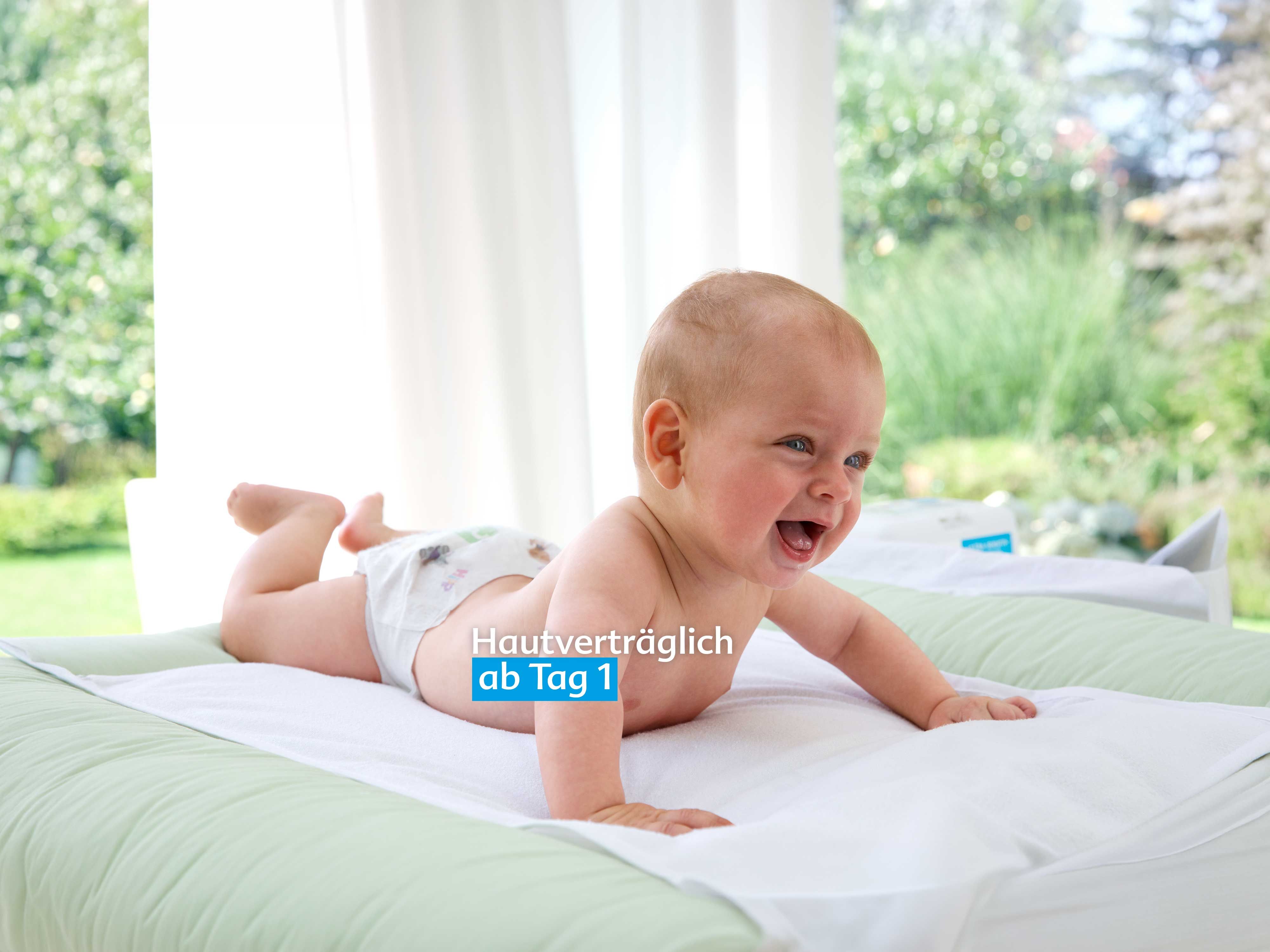 Lachendes Baby liegt auf dem Bauch auf einer Wickelunterlage vor einem Fenster mit Blick ins Grüne