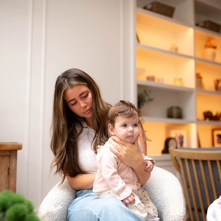A mother sitting in an armchair while holding her baby on her lap and gently supporting the baby's chin to burp them.
