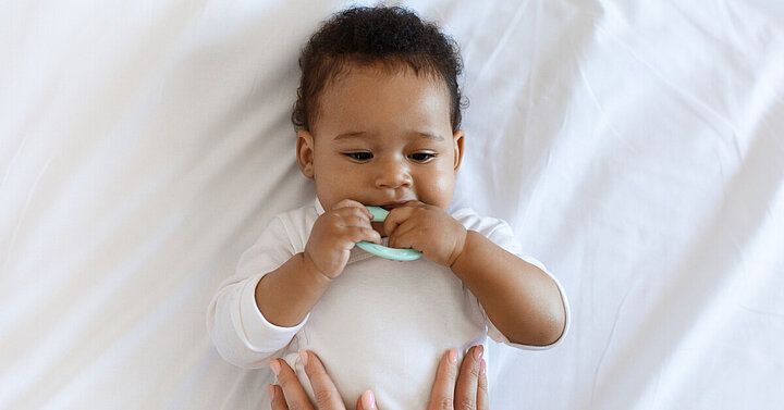 Smiling baby lying on a white bed, chewing a teething toy while a parent gently supports them