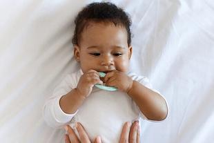 Smiling baby lying on a white bed, chewing a teething toy while a parent gently supports them