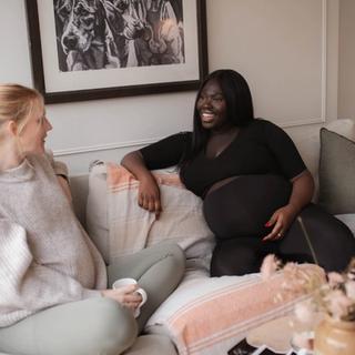 Two pregnant women sitting on a cozy couch, smiling and chatting with mugs in hand, surrounded by soft pillows and warm decor.