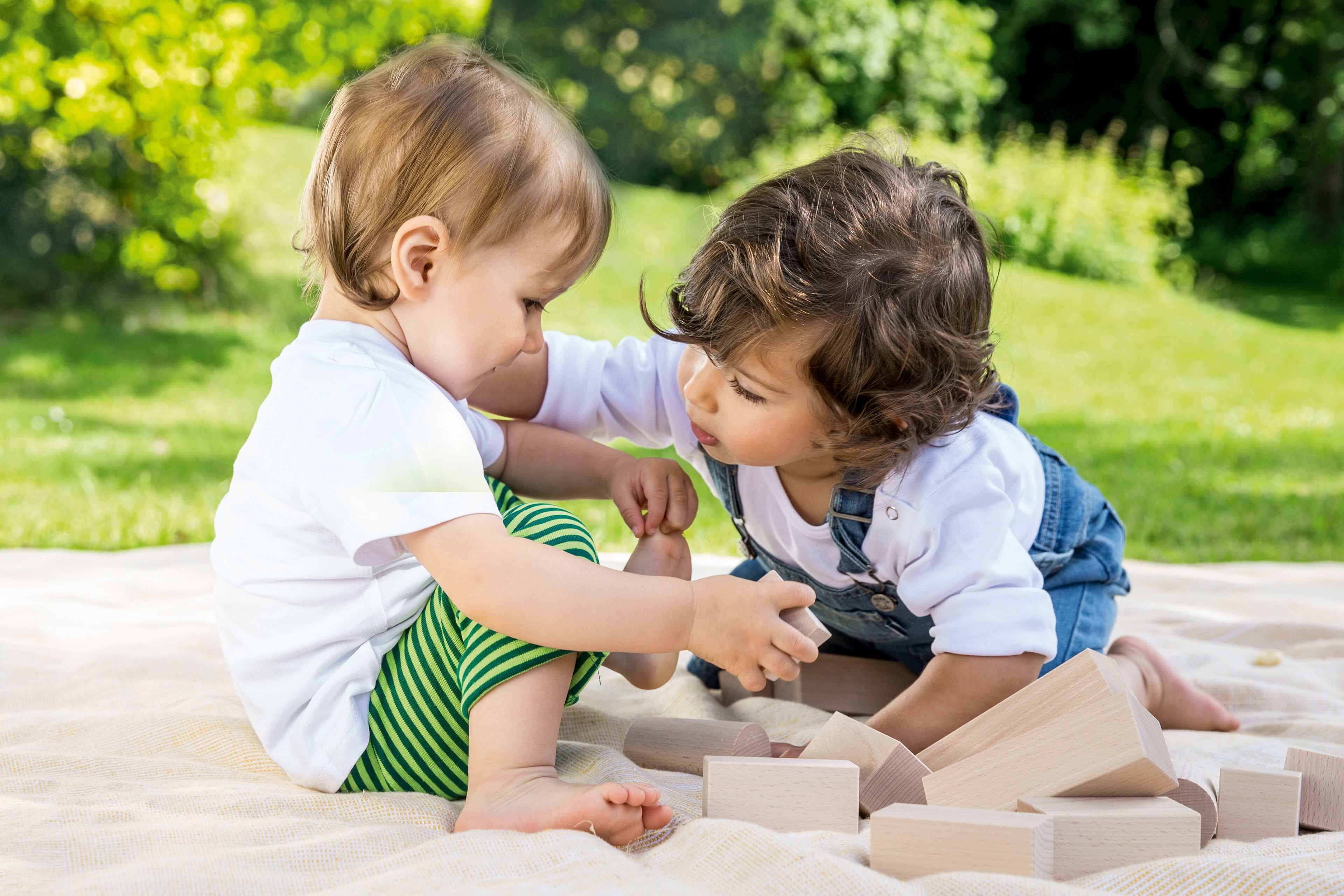 Zwei kleine Kinder, eines in einem weißen Hemd und das andere in einer blauen Latzhose, spielen mit Holzklötzen auf einer Decke im Freien.
