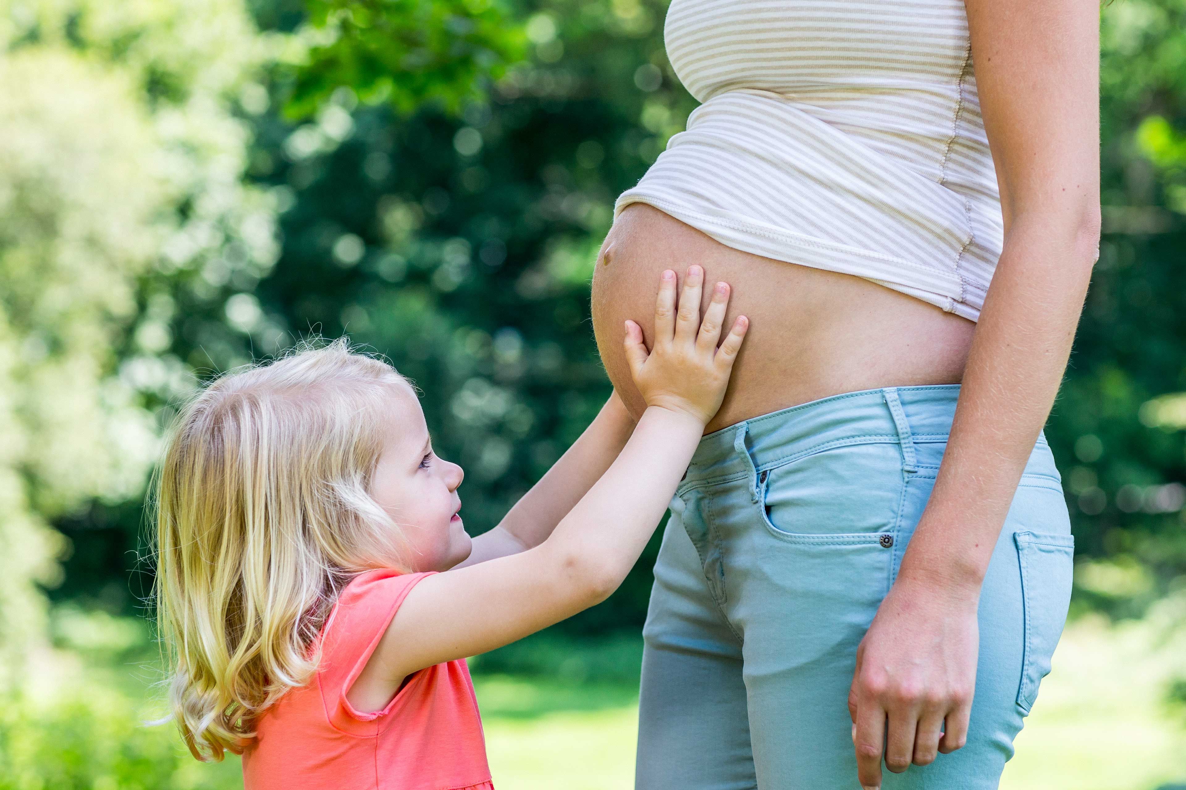 Ein junges Mädchen mit blondem Haar, das sanft den Bauch einer schwangeren Frau berührt, vor einem Hintergrund aus Grün.