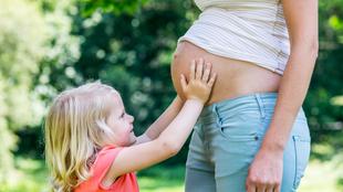 Ein junges Mädchen mit blondem Haar, das sanft den Bauch einer schwangeren Frau berührt, vor einem Hintergrund aus Grün.