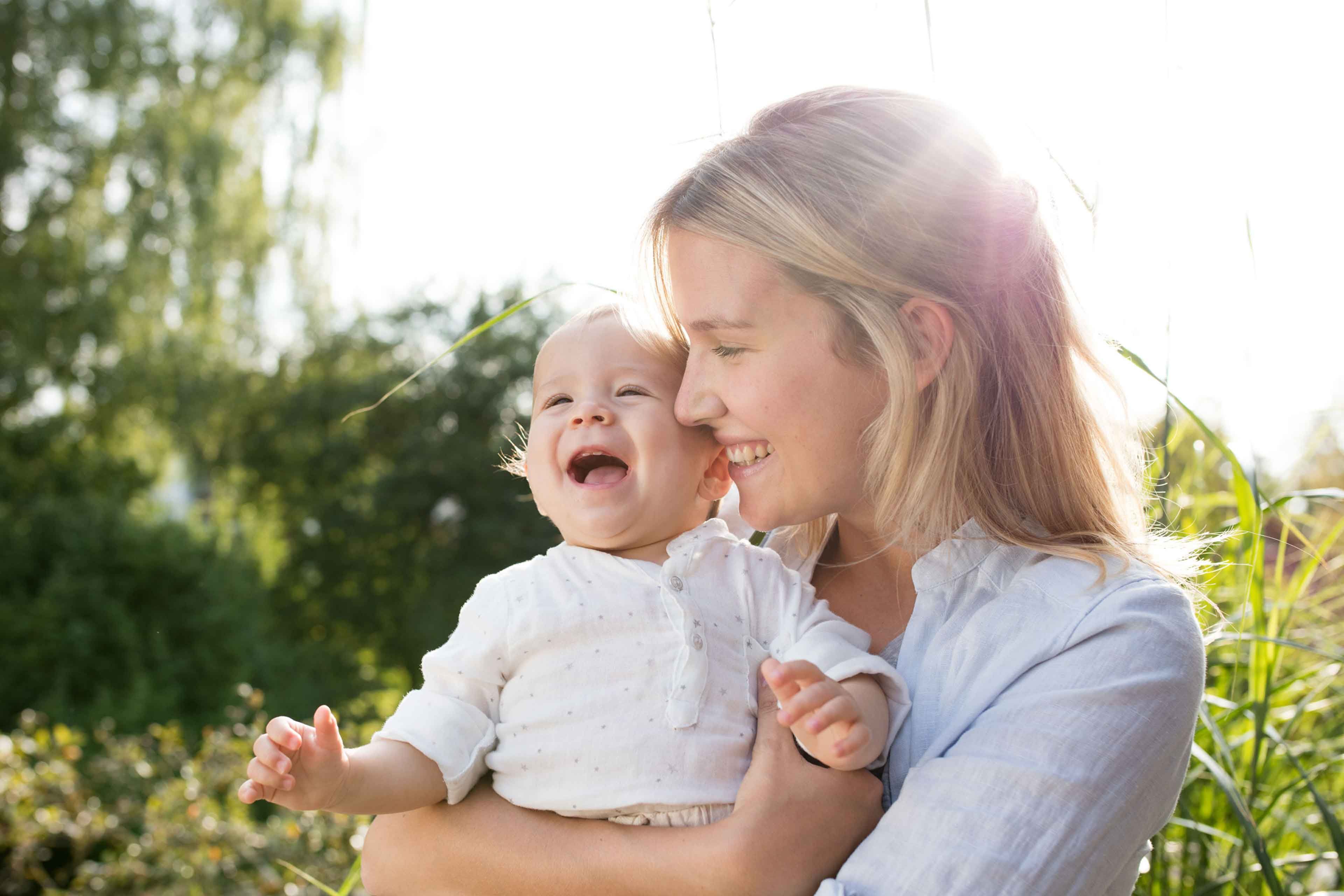 Lachende Mutter hält ihr fröhliches Baby im Grünen im Arm, Sonnenlicht im Hintergrund vermittelt Geborgenheit.