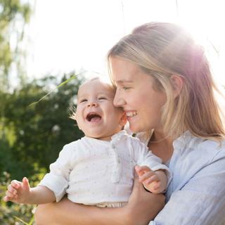 Lachende Mutter hält ihr fröhliches Baby im Grünen im Arm, Sonnenlicht im Hintergrund vermittelt Geborgenheit.