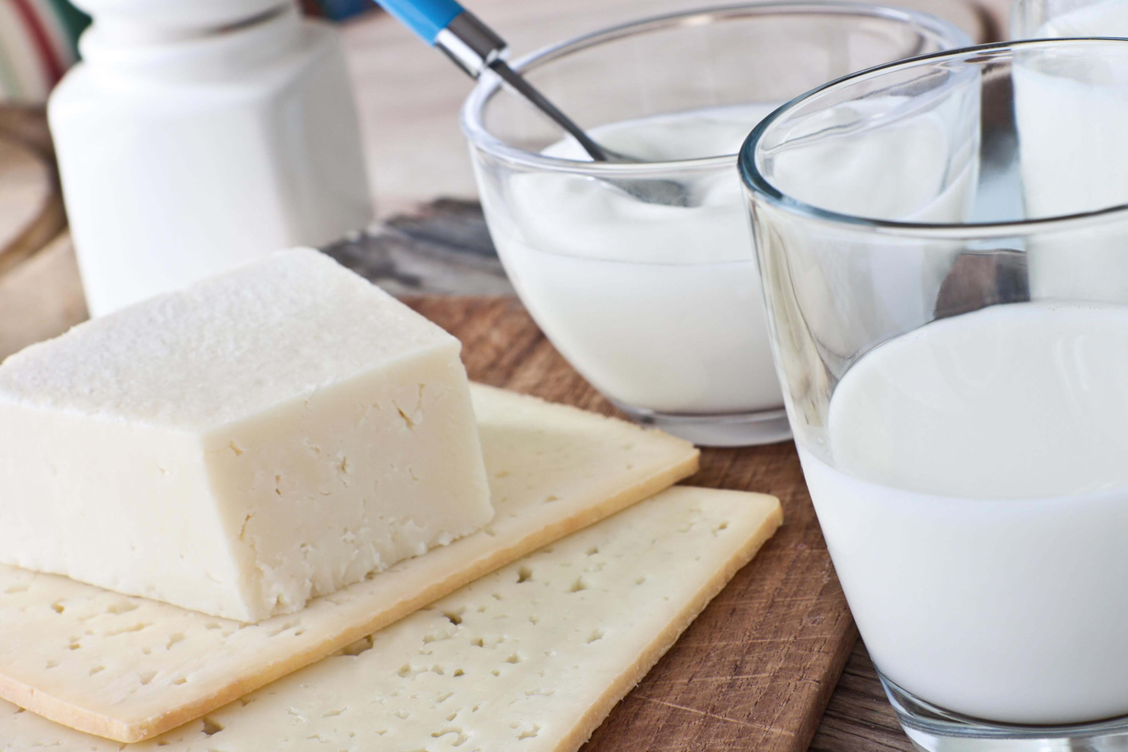Verschiedene Molkereiprodukte, darunter ein Stück Käse, ein Glas Milch und ein Behälter mit Joghurt, die alle auf einer Holzunterlage platziert sind.