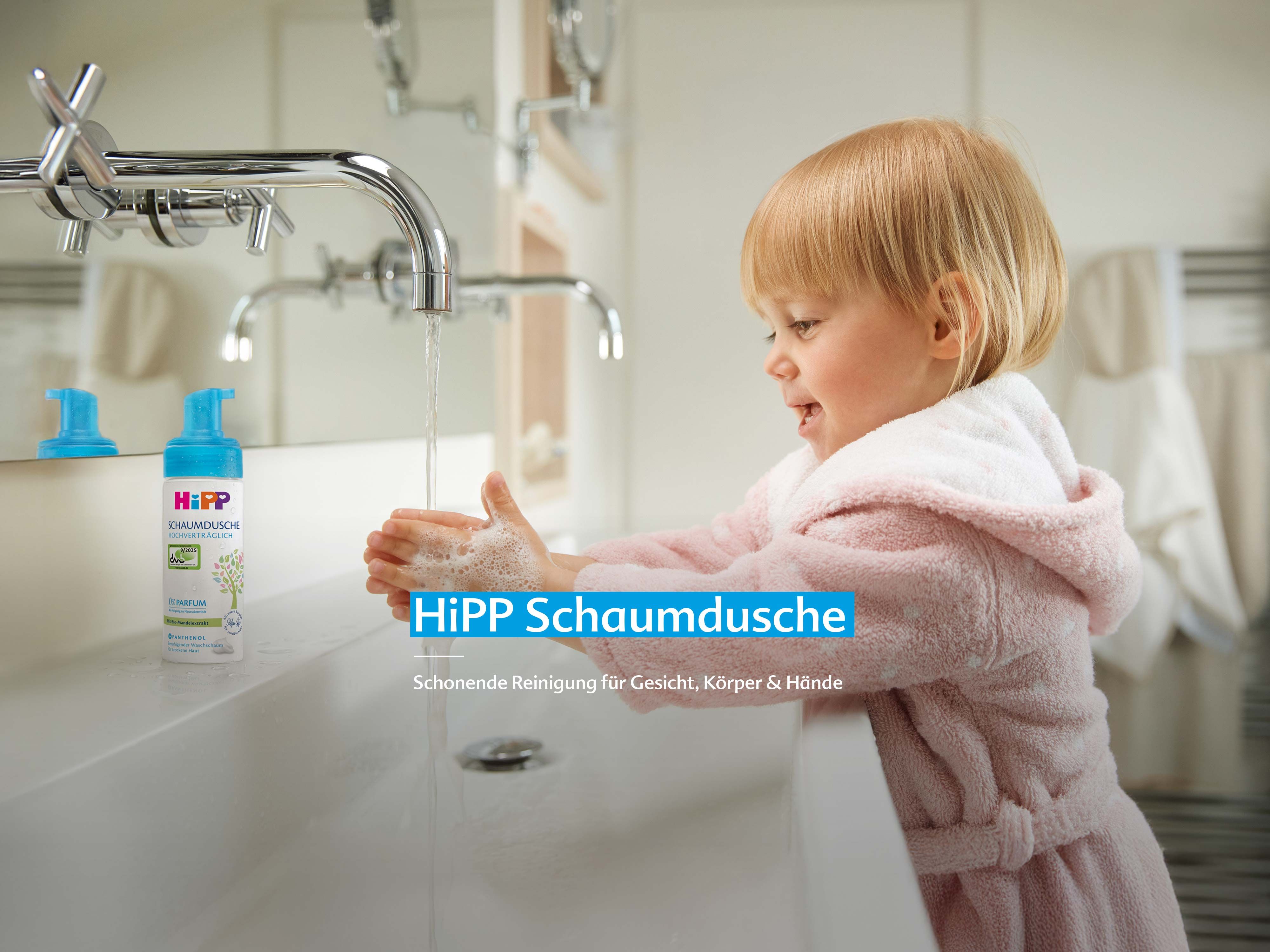 Kleines Kind im rosa Bademantel wäscht lachend die Hände mit HiPP Schaumdusche am Waschbecken mit fließendem Wasser.