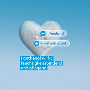 Herzförmiger Schaum vor blauem Hintergrund mit Beschriftungen 'Panthenol' und 'Bio-Mandelextrakt' sowie Text 'Panthenol wirkt feuchtigkeitsbindend und pflegend'