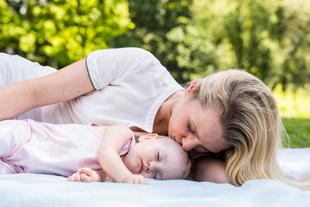 Mutter liegt im Grünen neben ihrem schlafenden Baby und küsst es sanft, während beide entspannt auf einer Decke ruhen.