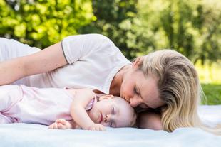 Mutter liegt im Grünen neben ihrem schlafenden Baby und küsst es sanft, während beide entspannt auf einer Decke ruhen.