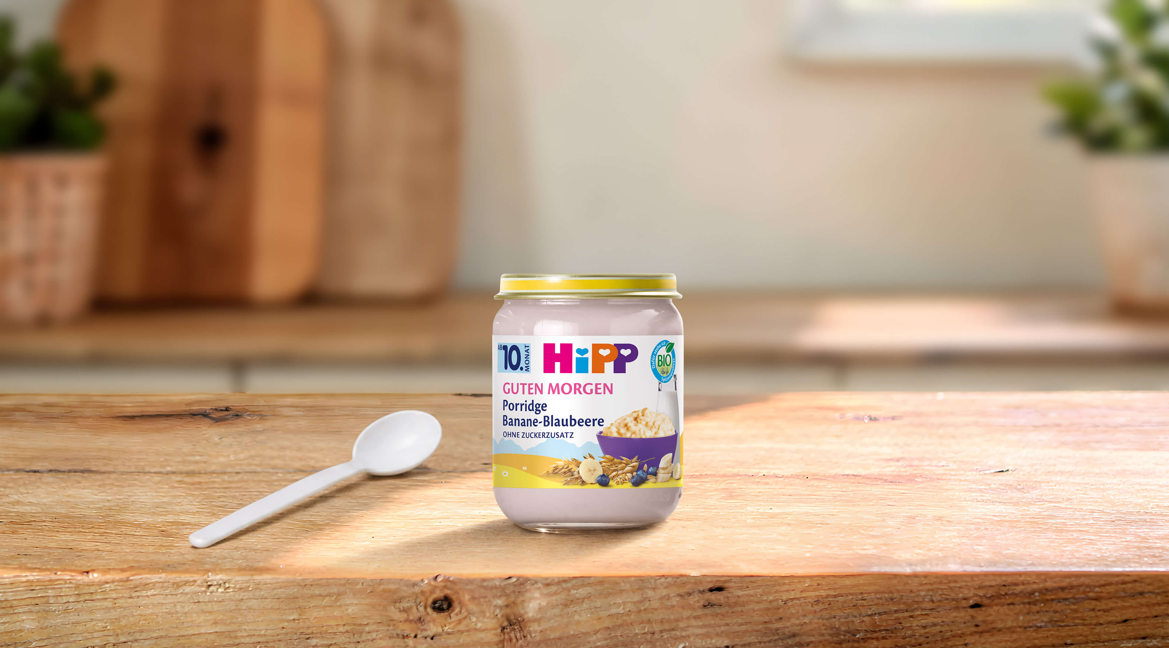 HiPP Guten Morgen Porridge Banane-Blaubeere im Glas mit Löffel auf einem Holztisch in heller Küche