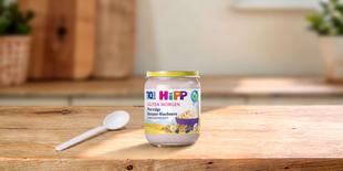 HiPP Guten Morgen Porridge Banane-Blaubeere im Glas mit Löffel auf einem Holztisch in heller Küche