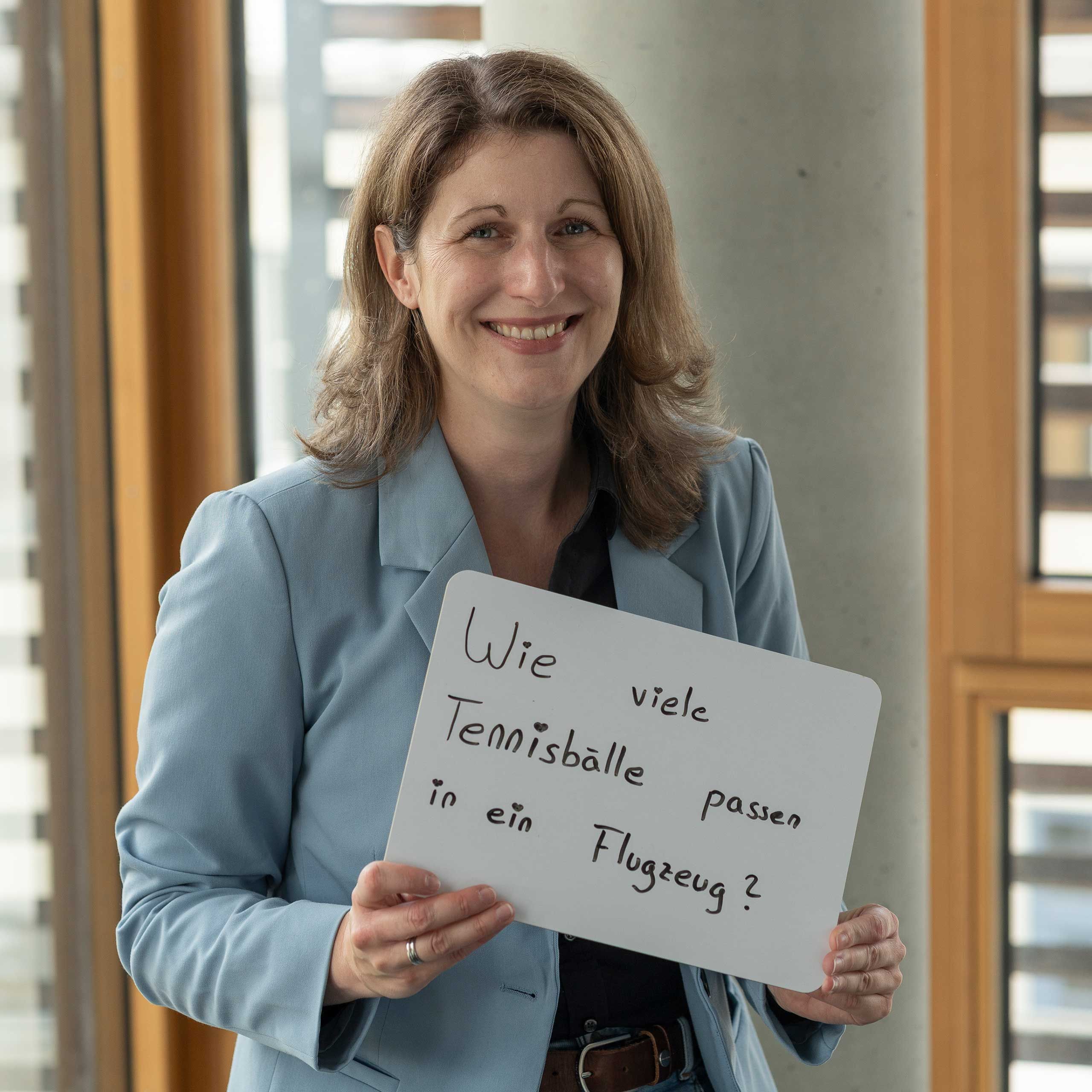 HiPP Mitarbeiterin hält ein weißes Schild mit der handschriftlichen Frage 'Wie viele Tennisbälle passen in ein Flugzeug'