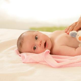 Baby liegt entspannt auf einem rosa Handtuch, wird sanft am Bauch gestreichelt und hält ein kleines Kuscheltier
