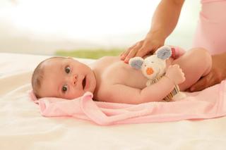 Baby liegt auf dem Rücken auf einer rosa Decke und hält ein Stofftier während eine Hand den Rücken massiert