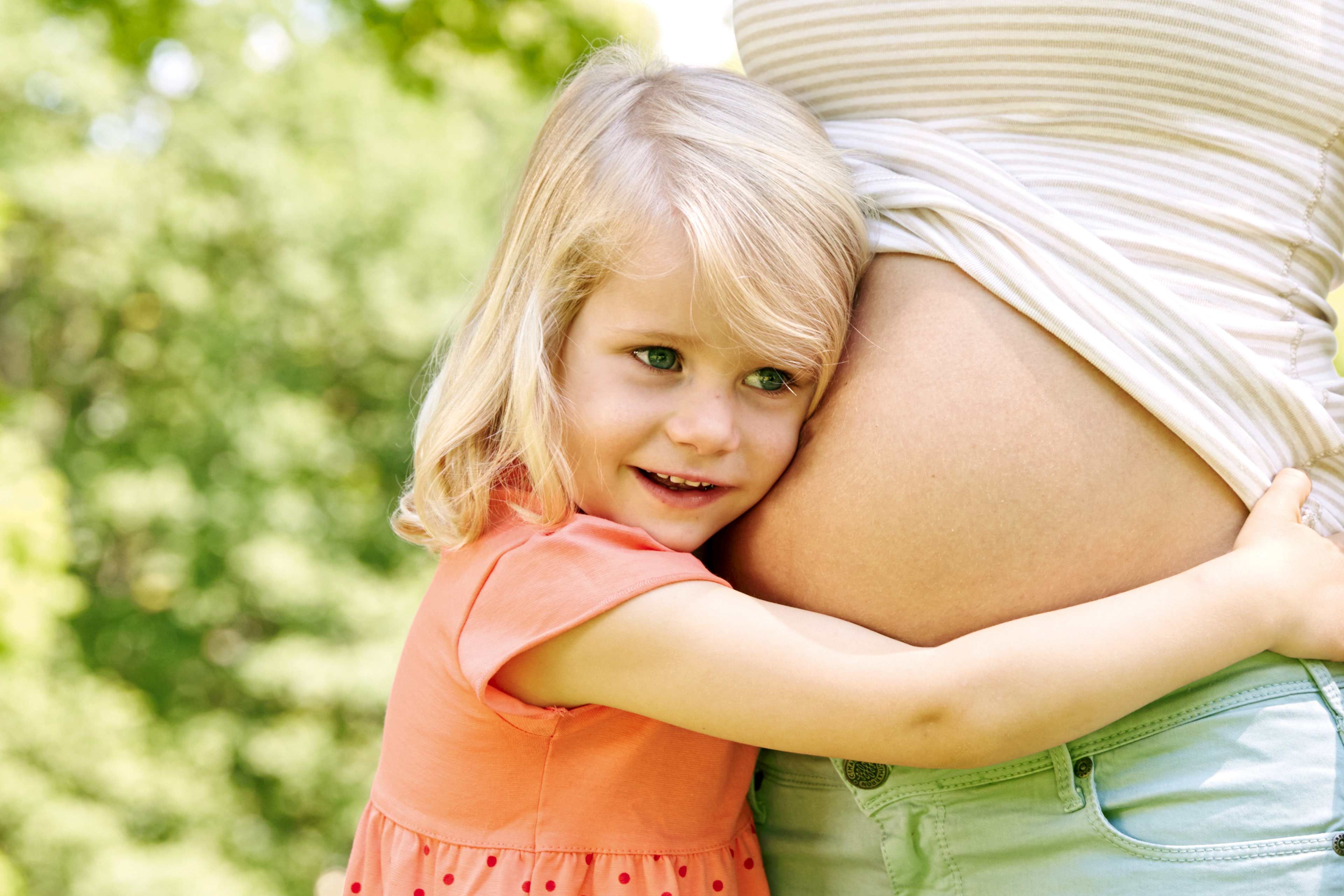 Ein junges Mädchen mit blondem Haar, das sich an den Bauch einer Frau schmiegt, vor einer grünen Kulisse.
