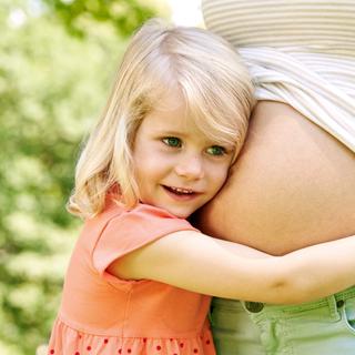 Ein junges Mädchen mit blondem Haar, das sich an den Bauch einer Frau schmiegt, vor einer grünen Kulisse.