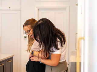 Two smiling women in a bright kitchen, one gently holding the other’s small baby bump in early pregnancy.