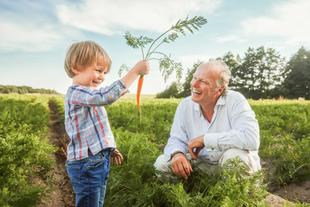 Kind hält eine frisch geerntete Möhre hoch und lächelt dabei, während Stefan Hipp in einem Feld hockt und ihn anlächelt