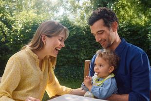 Vater und Mutter sitzen im Garten und schauen lächelnd auf ihr Kleinkind, das einen Löffel hält