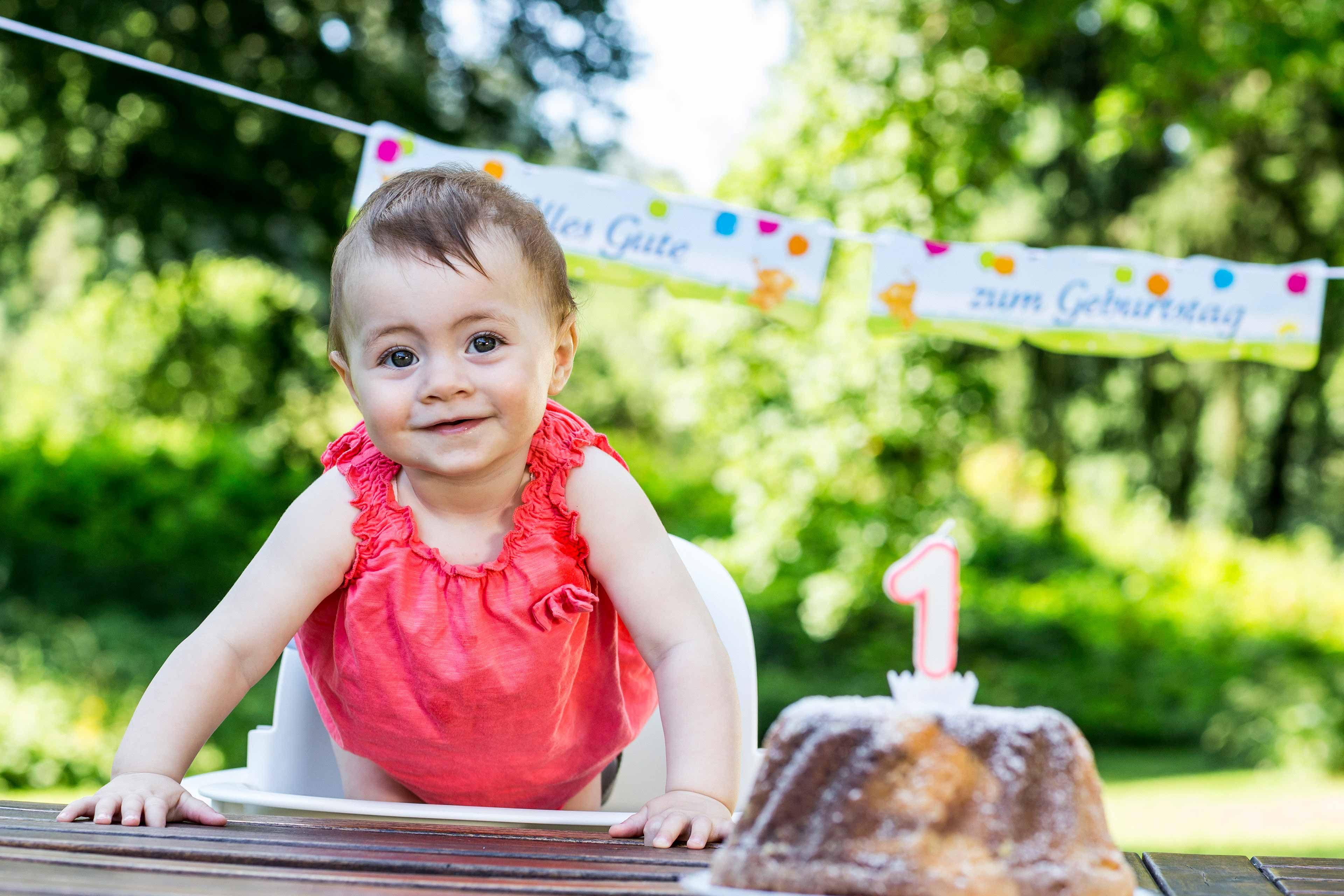 Ein kleines, rot gekleidetes Kind, das in einem Hochstuhl im Freien sitzt und vor sich einen mit einer einzigen Kerze verzierten Kuchen hat.