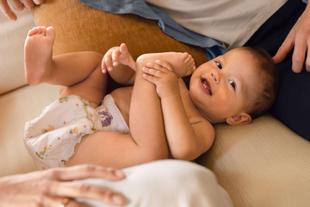 Lachendes Baby in Windel liegt entspannt auf dem Sofa und spielt mit den Füßen, umgeben von schützenden Elternhänden.