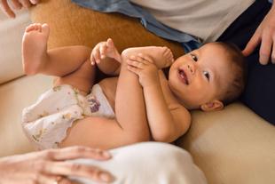Lachendes Baby in Windel liegt entspannt auf dem Sofa und spielt mit den Füßen, umgeben von schützenden Elternhänden.