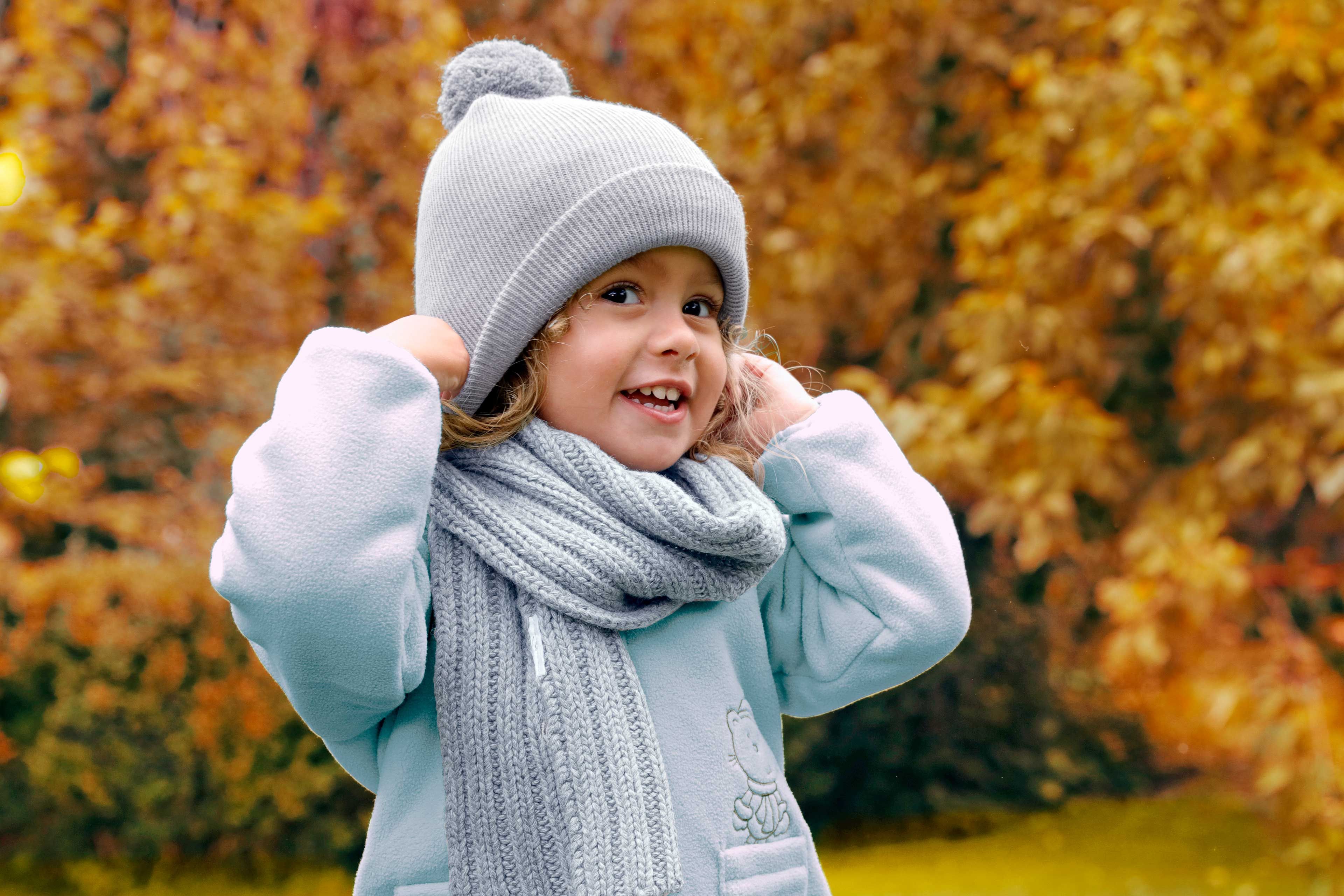 Kind mit Mütze und Schal hält sich die Mütze an beiden Seiten fest, herbstlicher Hintergrund mit Laub.