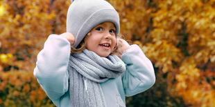 Kind mit Mütze und Schal hält sich die Mütze an beiden Seiten fest, herbstlicher Hintergrund mit Laub.