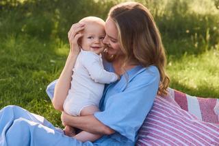 Frau im hellblauen Kleid sitzt auf einer Decke im Gras und hält ein Baby im weißen Strampler im Arm
