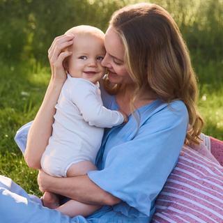 Frau mit hellblauen Kleid sitzt auf einer rosafarbenen Decke im Freien mit einem Baby auf dem Arm